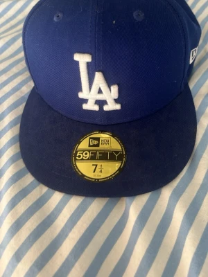 Blå LA Dodgers keps från New Era - Säljer en blå 59FIFTY keps från New Era med LA Dodgers-logga broderad i vitt framtill. Klassisk flat brim och officiell MLB on-field modell. Kepsen är i bomull och har detaljerade broderier samt New Era-logga på sidan.