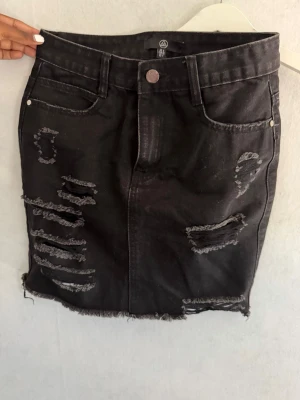 Svart jeanskjol med slitningar - Cool svart kort jeanskjol med slitna detaljer och rå kant nertill. Kjolen har fem fickor och stängs med knapp och dragkedja framtill. Perfekt för en trendig och avslappnad look.