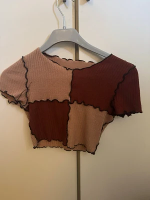 Patchwork croppad topp brun/beige - Trendig croppad topp i patchwork-stil med ribbad struktur. Toppen har korta ärmar och är sydd i block av mörkbrun och beige med vågiga svarta sömmar längs kanterna. Perfekt för dig som gillar en unik och edgy look.