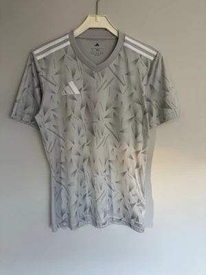 Grå mönstrad t-shirt från Adidas - Säljer en grå t-shirt från Adidas med coolt geometriskt mönster och vita detaljer. Tröjan har korta ärmar, v-ringning och klassiska Adidas-ränder på axlarna. Perfekt för träning eller sport. Materialet är lätt och andas bra.