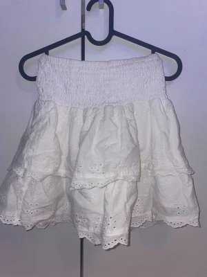 Vit volangkjol från H&M - Supersöt vit kortkjol från H&M med broderade detaljer och volanger. Kjolen har en smockad midja som gör den bekväm och lätt att bära. Perfekt för varma dagar och ger en fräsch och somrig look.
