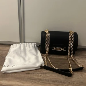 Svart axelväska från Michael Kors - Svart axelväska från Michael Kors i slät läderlook med guldfärgade kedjor och MK-detalj framtill. Väskan har en stilren, rundad form och levereras med en vit dustbag. Väskan har ett fack på baksidan och två inuti. Säljes då den tyvärr aldrig kommit till användning. 