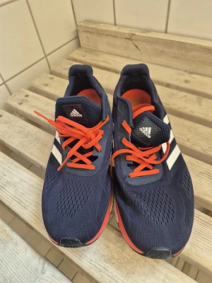 Adidas blå sneakers med röda detaljer - Snygga Adidas sneakers i marinblått med klassiska vita ränder på sidorna och röda snören samt röd sula. Ovandelen är i meshmaterial som andas och sulan är vit med bra dämpning. Perfekta för en sportig och stilren look.