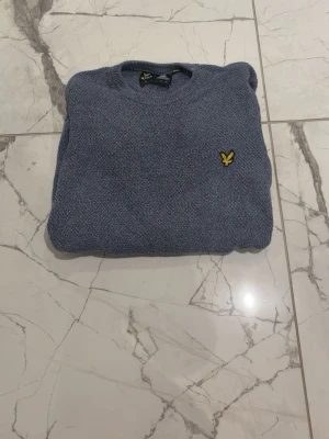 Blå stickad tröja från Lyle & Scott - Säljer en blå stickad tröja från Lyle & Scott med deras klassiska gula logga på bröstet. Tröjan har rund halsringning och långa ärmar. Perfekt för dig som vill ha en enkel men snygg look.