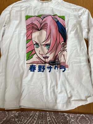 Vit Naruto långärmad t-shirt - Långärmad vit t-shirt med tryck av Sakura från Naruto på framsidan och blå Naruto-logga i nacken. T-shirten har en klassisk passform och är gjord i mjuk bomull. Perfekt för dig som gillar anime och vill ha en unik stil.