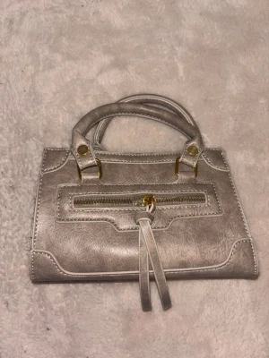 Liten grå handväska med gulddetaljer - Snygg liten handväska i grått konstläder med guldfärgade metalldetaljer. Väskan har dubbla handtag och en dekorativ dragkedja framtill med långa tofsar. Perfekt för dig som gillar stilrena accessoarer med en twist.