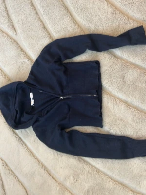 Mörkblå cropped hoodie med dragkedja - Säljer en mörkblå cropped hoodie från Bershka med dragkedja framtill. Hoodien har långa ärmar, ribbade muddar och en klassisk huva. Perfekt för en avslappnad och trendig look. Materialet är mjukt och stretchigt stickat tyg.