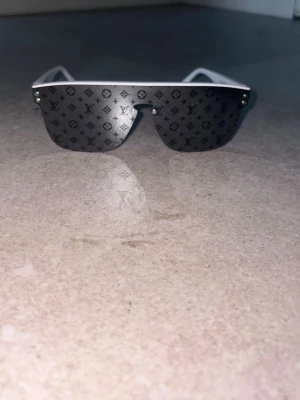 Vita solglasögon  -  Ett par moderna, kantlösa solglasögon med en futuristisk och exklusiv design. Glasen är mörkt tonade och har ett diskret monogrammönster över hela ytan. Fronten består av ett enda sammanhängande glas med en rak, vit överkant som ger ett stilrent och minimalistiskt uttryck. På sidorna finns små metallnitar som fungerar som dekorativa detaljer. Skalmarna är vita med en bred och robust konstruktion som förstärker den trendiga, statement-inspirerade looken.