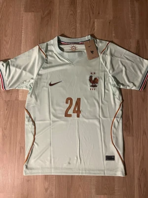 Cherki Frankrike VM-2026 Fotbollströja - Ljusgrön Frankrike fotbollströja med bronsfärgade detaljer och nummer 24. Tröjan har FFF-logga och tupp på bröstet, samt namn och nummer 'Cherki 24' på ryggen. Korta ärmar med randiga muddar och tunnt, ventilerande material. HELT NY