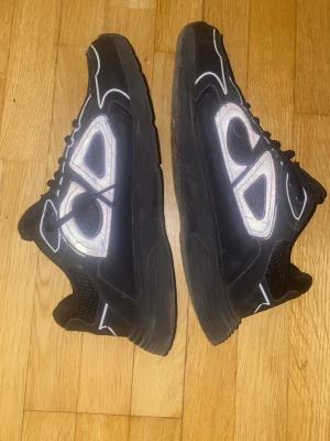 Svarta  sneakers med reflexdetaljer - Säljer ett par svarta sneakers med feta reflekterande detaljer på sidorna och vita linjer som kontrast. Skorna har en chunky sula och är tillverkade i en mix av mesh och syntetmaterial. Perfekta för dig som vill sticka ut med en lyxig streetwear vibe.
