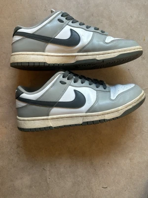 Nike Dunks - Säljer ett par Nike Dunk Low sneakers i grått och vitt med svart swoosh. Skorna har klassisk låg siluett, ovandel i läder och textilfoder. Yttersulan är i gummi och mellansulan är beige. Perfekta för dig som gillar retrostil och streetwear.