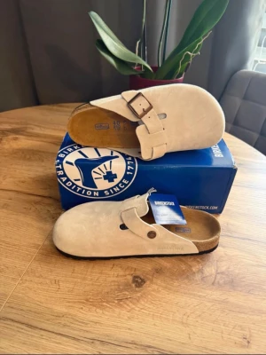 Beige Birkenstock Boston tofflor - Säljer ett par klassiska Birkenstock Boston tofflor i beige mocka med justerbart spänne i metall. Tofflorna har en ergonomisk fotbädd i kork och en mjuk ovandel i mocka för extra komfort. Perfekta för en avslappnad och stilren look.
