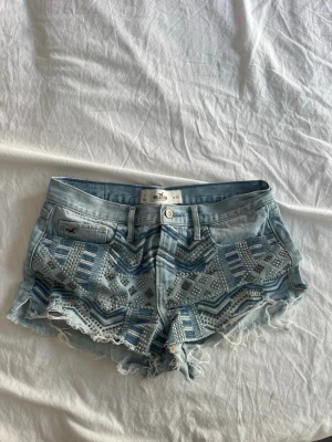 Ljusblå jeansshorts från Hollister - Säljer ett par ljusblå jeansshorts från Hollister med snygga slitna kanter och coolt mönster av nitar framtill. Shortsen har klassisk femficksmodell, knapp och dragkedja. Perfekta för sommaren och ger en avslappnad vibe.