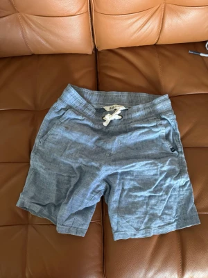 Blå linneshorts barn - Snygga blå shorts från H&M med vit snörning i midjan. Tillverkade i ett lätt och luftigt material, perfekta för varma dagar.  En liten fläck otvättad se sista bilden