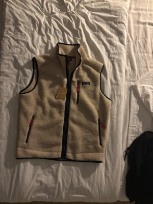 Beige fleeceväst från Patagonia - Säljer en beige fleeceväst från Patagonia med marinblå detaljer och dragkedjor med röda dragare. Västen har hög krage, tre fickor med dragkedja och är tillverkad i mjukt teddyfleece-material. Perfekt för lager-på-lager och outdoor-stil. Säljs då den inte passar mig, Kan ej garantera äktighet!! Därav kan pris diskuteras 