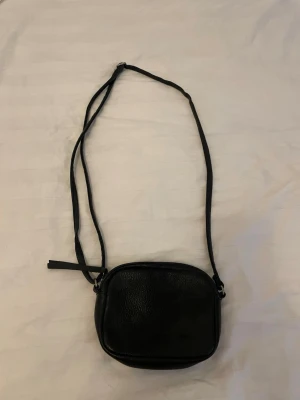 Svart liten axelväska i skinnimitation - Snygg svart axelväska i skinnimitation med justerbar rem och dragkedja upptill. Väskan är liten och smidig, perfekt för det viktigaste. Enkel design med rundade kanter och diskret struktur på ytan.
