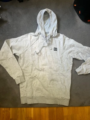 Grå hoodie från Under Armour - Snygg ljusgrå hoodie från Under Armour med klassisk logga på bröstet. Hoodien har huva med dragsko, känguruficka framtill och ribbade muddar vid ärmar och nederkant. Perfekt för chill dagar eller när du vill ha en sportig look. Passar perfekt till golfen vid frågor är de bara att skriver pris kan diskuteras (passar från 155-170 cm 