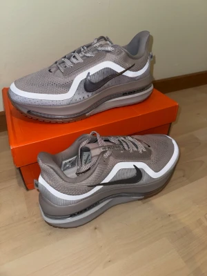 Nike Air Max Pegasus Zoom grå/vita sneakers - Nike Air Max Pegasus Zoom sneakers i grått och vitt med svart Swoosh. Ovandelen är i mesh och syntetmaterial för bra andningsförmåga. Tjock sula med synlig Air-enhet och vågiga detaljer längs sidan. Skosnören i matchande färg och klassisk Nike-design.