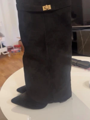 Lululia boots  - Snygga svarta höga boots i mocka med spetsig tå och dekorativt guldfärgat spänne upptill. Bootsens höga skaft ger en trendig siluett och materialet känns lyxigt. Perfekta för dig som vill ha en edgy och stilren look. Storlek 38