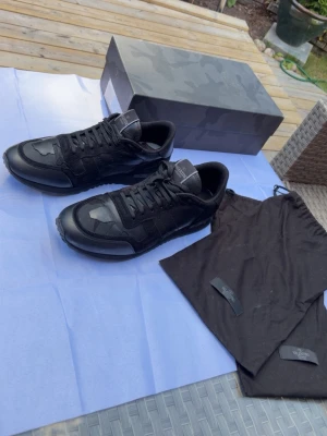 Svarta sneakers från Valentino - Snygga svarta sneakers från Valentino med coola camo-detaljer. Skorna har en mix av mocka och läder, samt svarta skosnören och diskret logga på plösen. Perfekta för dig som gillar stilrena men ändå unika sneakers. De är i fint skick men de är använda ett par gånger.