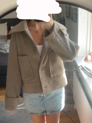 Beige kappa från Kappahl - Stilren beige jacka från Kappahl i ullmix med stora fickor framtill och bred krage. Jackan har knappar, uppvikta ärmslut och en croppad passform som ger en modern look. Perfekt för dig som vill ha en enkel men snygg jacka till vardags.