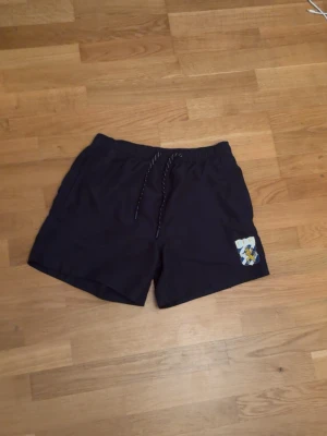 Ifk Göteborg badshorts - Säljer ett par mörkblå badbyxor i storlek S med resår och dragsko i midjan. På vänster ben finns ett färgglatt tryck med en tecknad figur. Badbyxorna har meshfoder på insidan och är perfekta för stranden eller poolen.