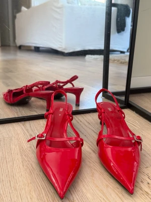 Röda lackpumps med remmar från Zara - Säljer ett par glansiga röda klackar från Zara med spetsig tå och smala remmar med spänne. Skorna har låg klack och öppen häl. Aldrig använt dessa men de är väldigt bekväma då har jag liknande i svart 