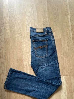 Blå jeans från Nudie Jeans - Säljer ett par klassiska blå jeans från Nudie Jeans med raka ben och femficksmodell. Jeansen har snygga kontrastsömmar samt Nudie-loggan broderad på bakfickan. Tillverkade i mjuk bomullsdenim som sitter skönt hela dagen. Jeansen har en lagning i skrevet som kan ses på 2 o 3 bilden, lagningen syns knappt när man har jeansen på sig 