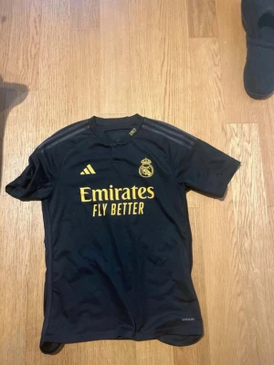 Real Madrid Bellingham matchtröja svart - Snygg svart Real Madrid matchtröja från Adidas med gula detaljer och tryck. Framsidan har klubbmärke och Emirates Fly Better-logga, baksidan har Bellingham och nummer 5 i gult. Tillverkad i lätt och ventilerande material, perfekt för fotboll eller träning.