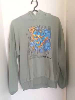 Grön hoodie - Ljusgrön hoodie från Lindex i storlek 158–164. Mjuk och bekväm modell med huva, avslappnad passform och snyggt grafiskt tryck fram. Använd men i fint skick utan hål eller större skador. Perfekt till vardags och enkel att matcha med jeans eller mjukisbyxor.