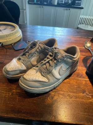 Nike Dunk Low gråvita sneakers - Nike Dunk Low sneakers i grått och vitt med klassisk Swoosh på sidan. Skorna har perforerad tåbox, platt sula och snörning. Tillverkade i läder och textil för en clean och tidlös look. De enda skadan på skorna är just baksidan, men går att laga på något sätt.