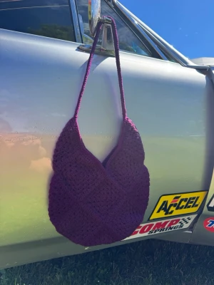 Lila virkad handväska - Unik handväska i lila, virkad för en trendig och personlig look. Väskan har en mjuk, rundad form och ett långt axelband som gör den enkel att bära. Perfekt accessoar för dig som vill sticka ut med en färgglad och handgjord stil. Material: akryl