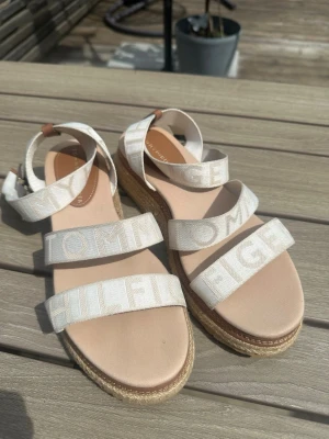 Beiga sandaler från Tommy Hilfiger - Snygga sandaler från Tommy Hilfiger med breda, vita textilband med logotyp. Sandalerna har en beige plattformsula i flätad jute och mjuk innersula. Perfekta för sommaren och ger en avslappnad men trendig look. Bara använt fåtal gånger och i väldigt fint skick.