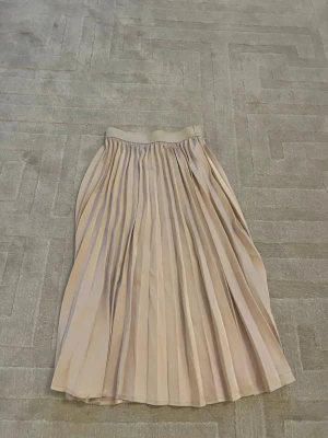 Plisserad beige långkjol i satin - Snygg plisserad långkjol i beige satin med bred resår i midjan. Kjolen har ett elegant fall och passar perfekt till en trendig och stilren look. Perfekt för dig som gillar en klassisk men modern stil.