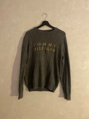 Grå kabelstickad tröja Tommy Hilfiger - Snygg grå kabelstickad tröja från Tommy Hilfiger med gul broderad logga på bröstet. Tröjan har rund halsringning, långa ärmar och ribbade muddar med diskreta rödvita detaljer. Perfekt för dig som gillar klassisk och stilren design.