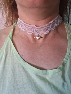 Spetschoker med pärldetalj och vinge - Säljer en vit spetschoker med hjärtformat mönster och en liten silverfärgad berlock i form av en vinge med en pärldetalj i mitten. Halsbandet har en romantisk och unik stil som sticker ut.