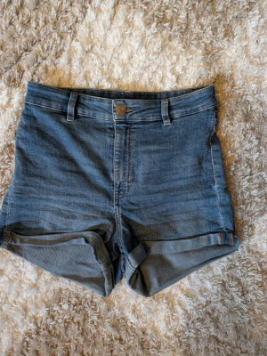 Blå jeansshorts med uppvik - Snygga blå jeansshorts med uppvikta benslut och klassisk femficksdesign. Shortsen har bälteshällor och stängs med knapp och dragkedja. Perfekta för varma dagar och enkla att matcha med olika toppar.