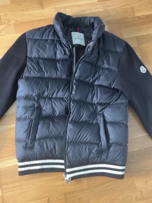 Svart cardigan från Moncler - Svart cardigan från Moncler med quiltad front och rygg, stickade svarta ärmar och ribbade muddar med vita ränder. Västen har två fickor med dragkedja och hög krage. Klassisk Moncler-logga på ärmen och innerfoder i blankt material. Storlek M men passar mest som S