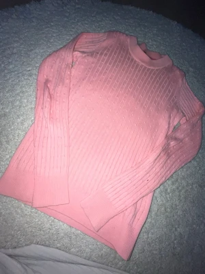Rosa ribbstickad tröja - Säljer en rosa ribbstickad tröja med rund hals och långa ärmar. Tröjan har ett diskret kabelstickat mönster och breda muddar vid ärmslut och nederkant. Perfekt för dig som gillar en enkel men snygg stil.