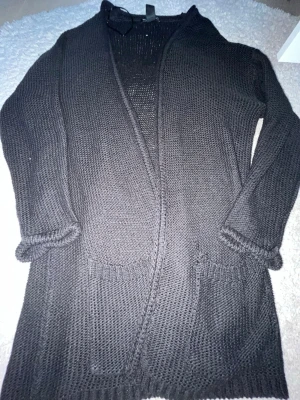 Svart stickad kofta med fickor - Mysig svart stickad kofta med öppen front och två praktiska fickor framtill. Koftan har långa ärmar och ribbstickade detaljer vid ärmslut och nederkant. Perfekt att slänga på sig när det är lite kyligt ute.