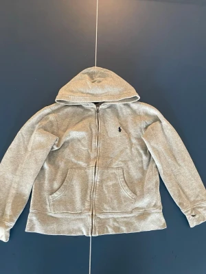 Grå hoodie från Polo Ralph Lauren - Snygg grå zip-hoodie från Polo Ralph Lauren med klassisk huva och två stora fickor framtill. Broderad logga på bröstet och mjukt, fleeceliknande innerfoder. Perfekt för en avslappnad och stilren look.