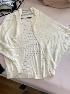 Vit öppen stickad kofta - Säljer en vit öppen stickad kofta med hålmönster och breda ärmar. Koftan har en avslappnad passform och ribbade kanter. Perfekt att slänga över en topp för en chill look.