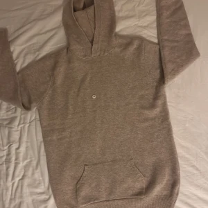 Beige merino hoodie  - Mysig beige hoodie med klassisk känguruficka och huva. Tröjan har en liten broderad logga på bröstet och ribbade muddar vid ärmslut och nederkant. Perfekt för en avslappnad stil.