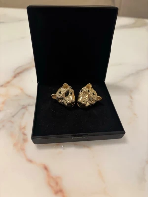 Guldörhängen leopardhuvuden - Unika örhängen i form av leopardhuvuden i guldfärgad metall med gnistrande stenar och svarta detaljer för ögon och nos. Djurinspirerad design som verkligen sticker ut och ger en cool touch till din stil. Perfekta för dig som gillar statement-smycken.