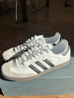 Adidas Samba - Klassiska Adidas Samba sneakers i vitt läder med svarta detaljer och de ikoniska tre ränderna på sidan. Skorna har vit ovandel, grå mockadetaljer vid tån och svart hälflik. Yttersulan är i gummi och snörningen är vit. Perfekta för en sportig och stilren look.