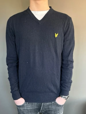 Mörkblå V-ringad tröja Lyle & Scott - Mörkblå V-ringad tröja från Lyle & Scott med den klassiska gula loggan broderad på bröstet. Tröjan har långa ärmar, ribbade muddar och är tillverkad i ett mjukt, tunt material som passar perfekt till jeans.      OBS! Modellen är 173cm 62kg