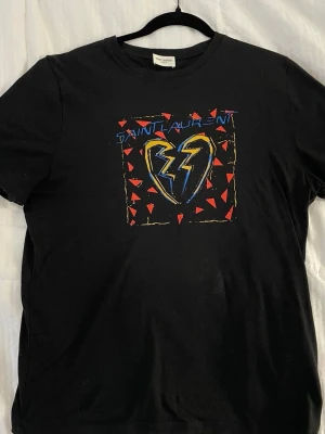 Svart Saint Laurent t-shirt - Cool svart t-shirt från Saint Laurent med färgstarkt tryck framtill som visar ett blixtformat hjärta och röda trianglar. Klassisk passform och rund halsringning. Perfekt för dig som vill sticka ut med en ikonisk design. ANVÄND TVÅ GÅNGER.