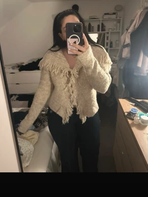 Beige kofta med fransar från Boohoo - Säljer en beige stickad kofta från Boohoo med stora fransar längs kragen och framkanten. Koftan har en bred krage och är tillverkad i ett mjukt, ull-liknande material. Perfekt för dig som gillar en unik och cozy look.