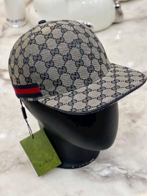 Gucci beige keps med GG-mönster - Snygg beige keps från Gucci med klassiskt GG-monogram över hela kepsen. På sidan finns en röd och marinblå rand som ger extra stilpoäng. Kepsen är tillverkad i canvasmaterial och har en böjd skärm. Perfekt för dig som vill ha en exklusiv och trendig accessoar.