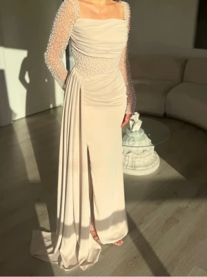 Elegant beige långklänning med pärlor - Säljer en elegant beige långklänning med pElegant beige långklänning med pärlorärlbroderade långärmar och detaljerad midja. Klänningen har draperad front och slits, vilket ger en lyxig känsla. Perfekt för dig som vill sticka ut med en unik och stilren look.